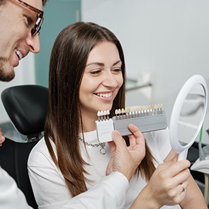 Woman in dental chair smiling into mirror as dentist holds shade guide to her grin