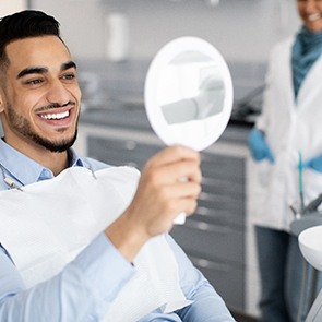 Man with dark hair in dental chair smiling into mirror with dentist blurry in background